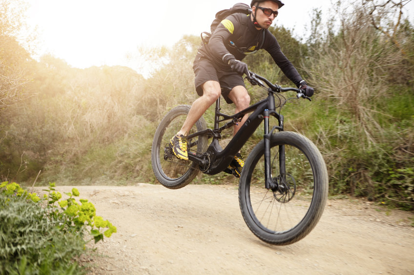 Cyclist in action jumping high on black electric motor powered bicycle down trail in woods. Young rider wearing glasses and helmet making extreme biking stunt on e bike while exercising outdoors